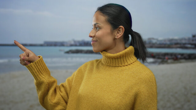Woman in yellow sweater points at seaside on beach with cloudy sky, showcasing serene and calm coastal environment while smiling peacefully outdoors with ocean background. - Powered by Adobe