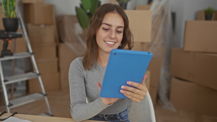 Young woman smiling while holding blue tablet and tapping touchscreen amid packed moving boxes in house; new beginnings optimism.