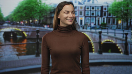 Woman smiling with face visible on street by amsterdam canal bridge, wearing brown turtleneck; serenity.