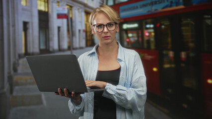 Blond woman wearing glasses and a light shirt holds a laptop and types on the keyboard with visible hands on a busy city street beside a red doubledecker bus; urban work concentration.