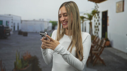 Young woman texting on smartphone with clenched fist on a sunlit street near outdoor chairs and potted plants; joy success.