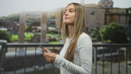 Young hispanic woman holding smartphone, typing with both hands on a balcony of a historic stone building overlooking archaeological ruins; urban confidence.