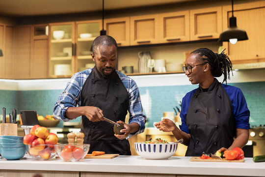 Smiling boyfriend cutting avocado for a fresh summer salad in the kitchen, preparing a healthy lunch meal with organic vegetables as ingredients. African american couple chopping veggies. - Powered by Adobe
