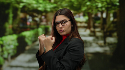 Confident hispanic woman in glasses with clenched fists stands determinedly in a vibrant park setting, surrounded by lush greenery and a sunlit pathway ahead.