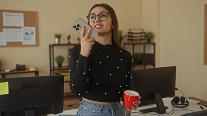 Young woman in an office holding a phone and red mug, wearing glasses, speaking in a modern workplace with computers and documents around, conveying multitasking productivity.