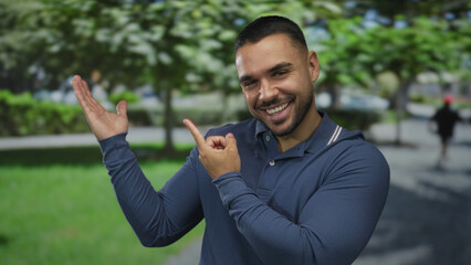 Young hispanic man smiling broadly and pointing finger to his left in street park under green...