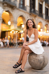 Obraz premium Evening Royal square in Barcelona, girl in sundress is sitting on stone ball, resting on concrete fencing. Electric street lighting, twilight in southern city, evening life in big city