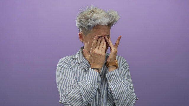 Woman with short hair covers eyes with hands in purple studio set; sadness isolation grief despair.