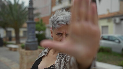 Woman with focused gaze and outstretched hand raised to block the camera on a sunlit urban street with palm trees and a car in view; defiance.
