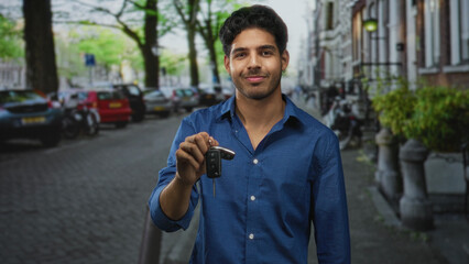 Man extending hand holding car keys, smiling with eyes closed, wearing blue shirt on street; joy pride.
