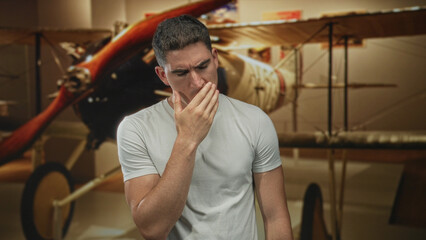 Young hispanic man grimacing and clutching chest beside vintage biplane in museum building exhibit  heartattack pain. © Krakenimages.com