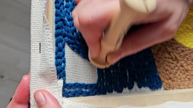A person uses a hook to craft a rug with blue, green, and yellow colors. The setting has a soft texture and features detailed patterns while taking place indoors.