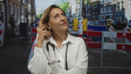 Hispanic woman doctor wearing white coat and stethoscope touches hair while gazing upward on street...