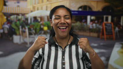 Obraz premium Woman referee clenches fist in busy outdoor street soccer game in front of cafe; celebration joy victory.