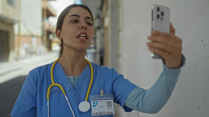 Young hispanic woman doctor wearing uniform and stethoscope holds smartphone for video call on street; telemedicine remote care empathy.
