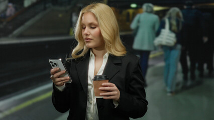 Woman browsing phone in stylish suit while holding coffee at busy underground station platform with...