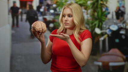 Young woman holding model brain outdoors on busy city street reflecting confusion and curiosity with blurred urban background contrasting her casual red shirt presence