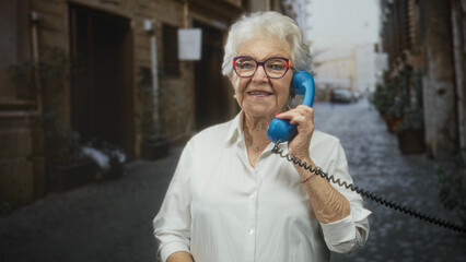 Elderly woman holds blue phone handset to ear while making ok gesture on cobblestone street;...