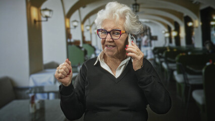 Elderly woman holds phone to ear and makes a fist gesture inside a building cafe with arched seating and lamps; joy connection.