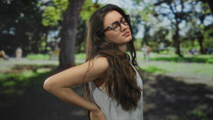 Woman with glasses holds lower back showing backache in forest clearing while wearing white sleeveless top; discomfort.