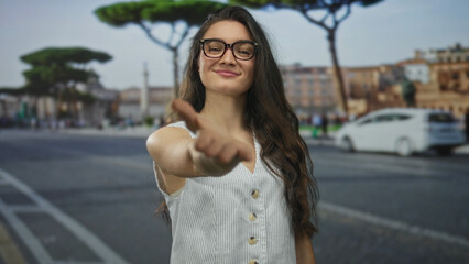 Fototapeta premium Young hispanic woman with glasses extends open hand on cobblestone street in white button dress; generosity.