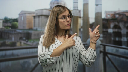 Blonde woman finger pointing to building facade under cloudy daylight on outdoor balcony railing by historic ruins; curiosity.