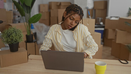 Woman working on laptop with phone held to ear amid stacked moving boxes inside a house building, typing on keyboard; focused small business planning.