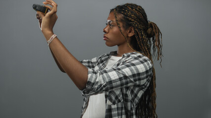 Woman with braided hair holding a compact camera aloft, wrist and forearm visible, wearing plaid shirt in a grey studio; concentration.