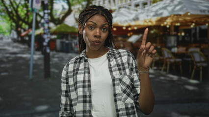 Woman with braided hair wearing white tee and plaid shirt points index finger up on restaurant terrace under string lights; confidence reminder.