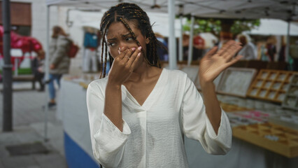 Woman pinching nose and covering mouth at a busy outdoor market stall with souvenir tables, reacting to a strong smell in the street; disgust concern.