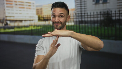 Young man gestures time out on street in urban city with smile wearing white shirt outdoors under...