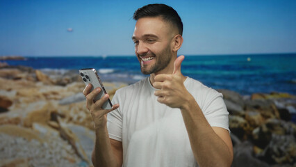 Young man smiling at beach while using smartphone and giving thumbs up by the sea.