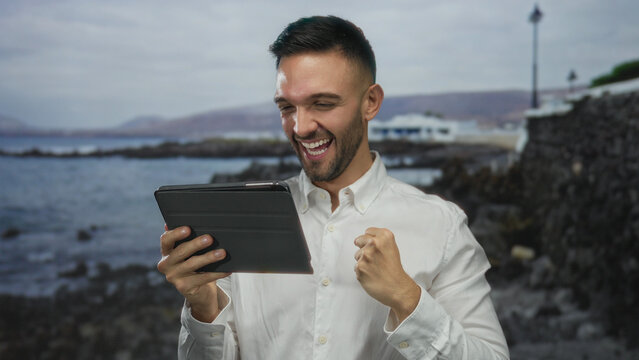 Young man with tablet smiling on a beach with a scenic seashore in the background showcasing technology and travel outdoors in a vibrant coastal setting.