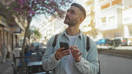 Young man outdoors using smartphone on sunny day, wearing casual clothing, with urban background, focusing on screen, surrounded by blooming trees and buildings in bright setting.