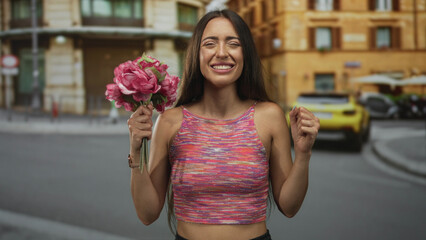 Fototapeta premium Woman holding bouquet and raising fist in street near parked car and stone building; joy celebration.