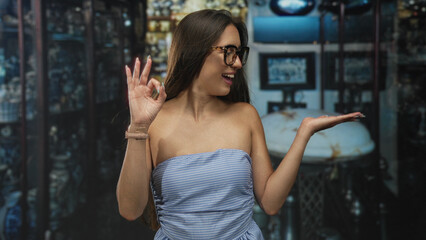 Woman wearing glasses and a strapless dress showing ok sign with bare shoulders and open smile in studio; confidence.