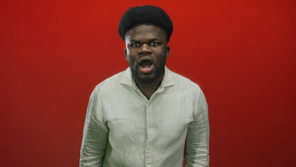 Obraz premium Young african american man shouting with open mouth and intense gaze wearing light linen shirt in studio; anger defiance protest.