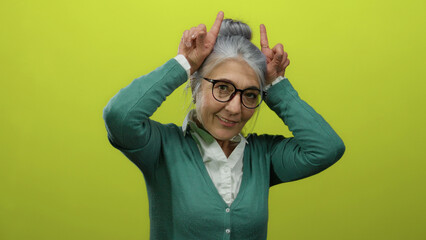 Senior woman with glasses in green cardigan having fun gesturing with fingers as horns against...