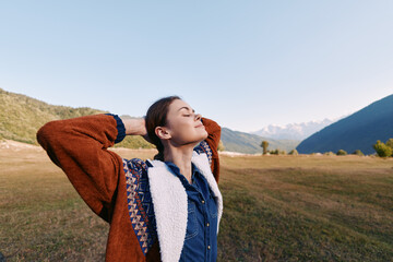 Fototapeta premium Woman in nature among mountains and meadow, portrait outdoors to relax wearing a warm sweater, eyes closed and breathing deeply while enjoying peaceful rural scenery and sunlight.