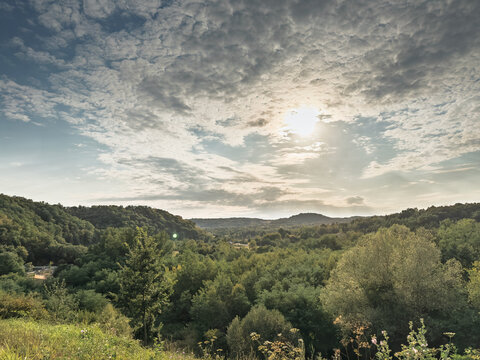 Wide panorama of the hills of Sumadija in central Serbia. Sunlight breaks through clouds above a mosaic of forests, meadows and valleys, illustrating the region's green rural landscape.