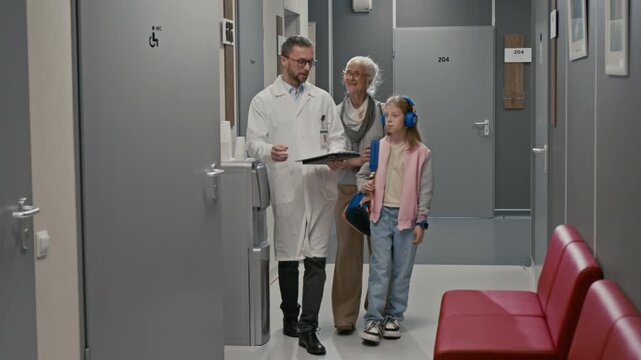 Front view shot of smiling senior woman and child following doctor and walking towards camera while discussing recommendations in clinic hall, copy space