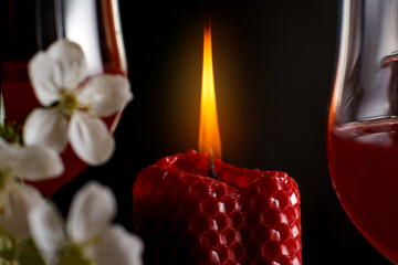 Close up of a red candle with a bright flame and white flowers and a glass of red liquid nearby