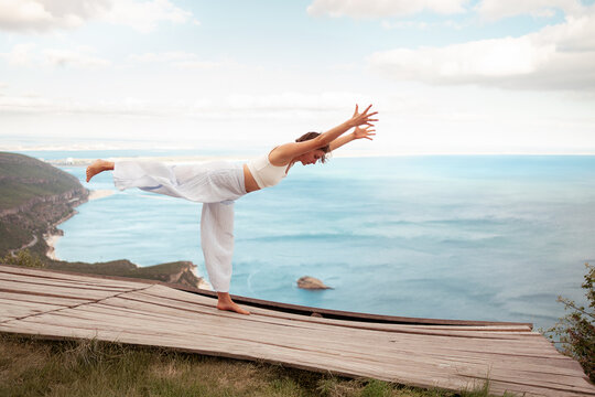 Full body of woman practicing Warrior III Pose yoga on wooden platform by ocean. Fit lady stretches gracefully representing strength and serenity in peaceful scenic nature