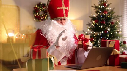 Saint Nicholas having video chat via laptop at desk in room decorated for Christmas. Camera moving forward