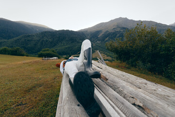 Fototapeta premium Person on bench in mountains nature relax boots meadow outdoor lying on wooden plank with jacket, peaceful landscape, distant peaks and green valley for weekend escape and rest.