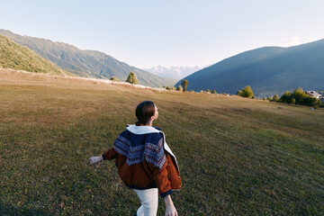 Fototapeta premium Woman walking in a meadow toward mountains across open landscape, nature hiking outdoors travel scene with jacket and shawl, back view capturing freedom and scenic valley peace.
