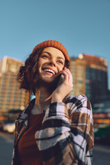 Fototapeta premium Woman on phone smiling in city with beanie and flannel, earbuds in sunlight. Candid lifestyle portrait at golden hour glow, authenticity and mindful living with emotional storytelling.