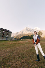 Fototapeta premium Woman in a jacket and boots standing in a meadow near a rustic building with alpine mountain backdrop, outdoor portrait capturing scenic landscape, sky and peaceful countryside view.