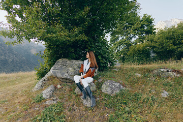Fototapeta premium Woman sitting on rock under tree in mountain meadow outdoors, portrait of a traveler in boots and warm coat enjoying the landscape and peaceful autumn nature with distant peaks.