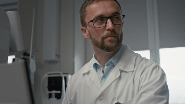 Tilt up shot of male doctor as optometrist using modern equipment in clinic and pressing buttons on keyboard with focus flowing from hands to face, copy space